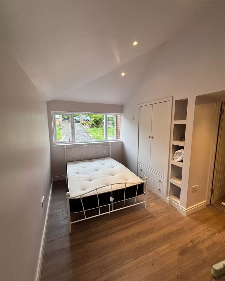 Garage converted into a bedroom by SMM — vaulted ceiling with downlights, built-in wardrobe and shelving, and a wide window